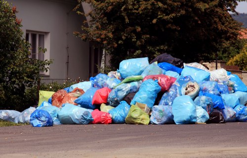 A large outdoor pile of mixed rubbish bags and loose waste materials situated on the edge of a paved driveway in a residential area. The accumulation includes numerous plastic bags in various colours, primarily blue, with some red, green, orange, and black bags. The bags appear to be filled with household waste, and some may contain visible debris such as paper, plastic, and other refuse. To the left, there is part of a white building with a window, and behind the pile, there is a large leafy tree providing partial shade. The background features additional greenery and possibly parts of other residential structures, suggesting the waste is temporarily stored before collection. The scene is indicative of waste clearance work that a professional rubbish removal service, such as Business Waste Removal Sydenham, might manage for local homeowners or small businesses in the area, aligning with their services for efficient rubbish disposal in the local SE26 postcode district.