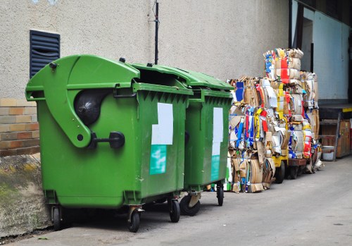 A row of three large green wheelie bins with black lids, positioned on a paved driveway adjacent to a beige exterior wall of a building. The bins are made of durable plastic with textured surfaces and are equipped with small wheels at the base for mobility. Two of the bins have white labels attached to their fronts, indicating recycling or waste types. To the right of the bins, there are multiple tightly packed bundles of flattened cardboard and paper materials, stacked against a wooden pallet. The bundles display various colours and textures of cardboard packaging, including brown Kraft, white, and blue printed surfaces. The scene is situated in an urban or suburban setting, with a concrete pavement and a small portion of a brick wall visible at the lower left. The background includes a partially visible window and door of the building, with diffuse natural lighting suggesting a typical daytime environment. This image relates to rubbish collection services provided by Business Waste Removal Sydenham, serving the local area, including local postcode conjecture, for efficient waste and recycling removal.