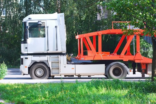 A white flatbed truck with a grey cab is parked on a paved road with a grassy verge in the foreground. The truck has a red metal frame structure on its bed, designed for carrying and lifting bulky waste or construction debris. The red frame includes vertical supports and a curved railing around the edges, providing stability and safety for loaded items. The background features a line of dense green trees and shrubbery, typical of a suburban or semi-rural environment in Sydenham, London postcode SE26. The area appears outdoors during daylight, with natural light illuminating the scene evenly. Business Waste Removal Sydenham utilizes such trucks for rubbish and waste collection services, often seen operational in local residential or commercial areas to facilitate efficient waste management and clearance. The scene is clean and orderly, emphasizing practical transport equipment used for rubbish removal and disposal tasks consistent with local waste management requirements.