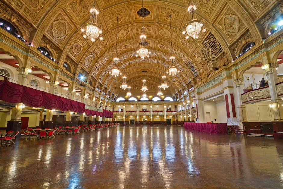 The image depicts a large, ornate interior space resembling a historic ballroom or event hall, characterized by a high, intricately decorated vaulted ceiling with detailed moldings and multiple hangin