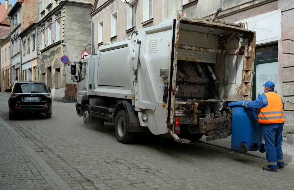 A compact light blue flatbed truck parked on the side of a street, overloaded with a large amount of mixed waste materials including cardboard boxes, black plastic bags, and bundled items, all piled h